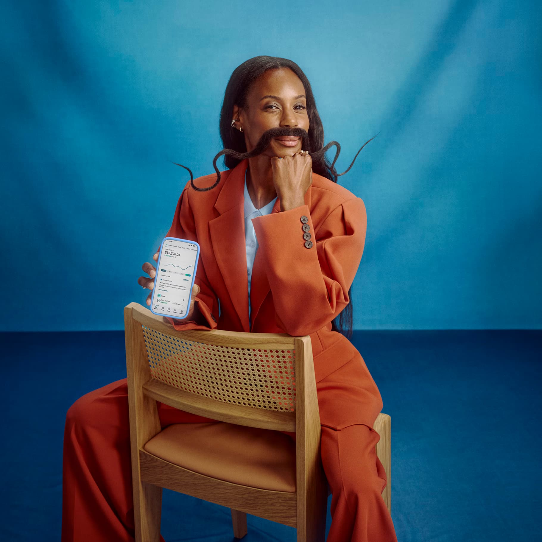 A woman in an orange suit with a long curled moustache sits on a chair, holding up a phone showing the Stash app
