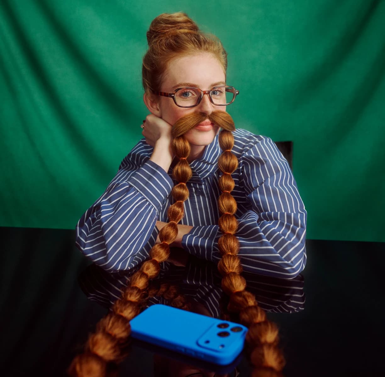 A woman in glasses and a striped shirt with a long braided moustache, a blue phone on the table in front of her